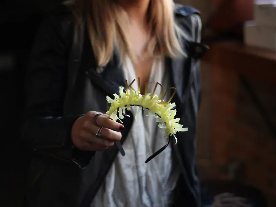 A hand holding a headband trimmed with pale yellow flowers against a dark jacket