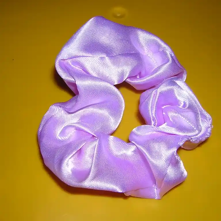 A gathered lilac satin scrunchie lying on a mustard-yellow ground